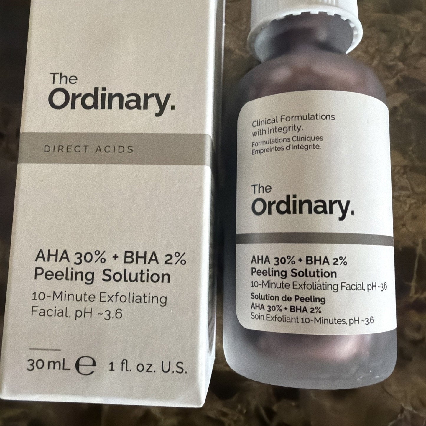 Box and frosted glass bottle of The Ordinary AHA 30% + BHA 2% Peeling Solution with minimalist black text on a white background, placed on a textured surface.