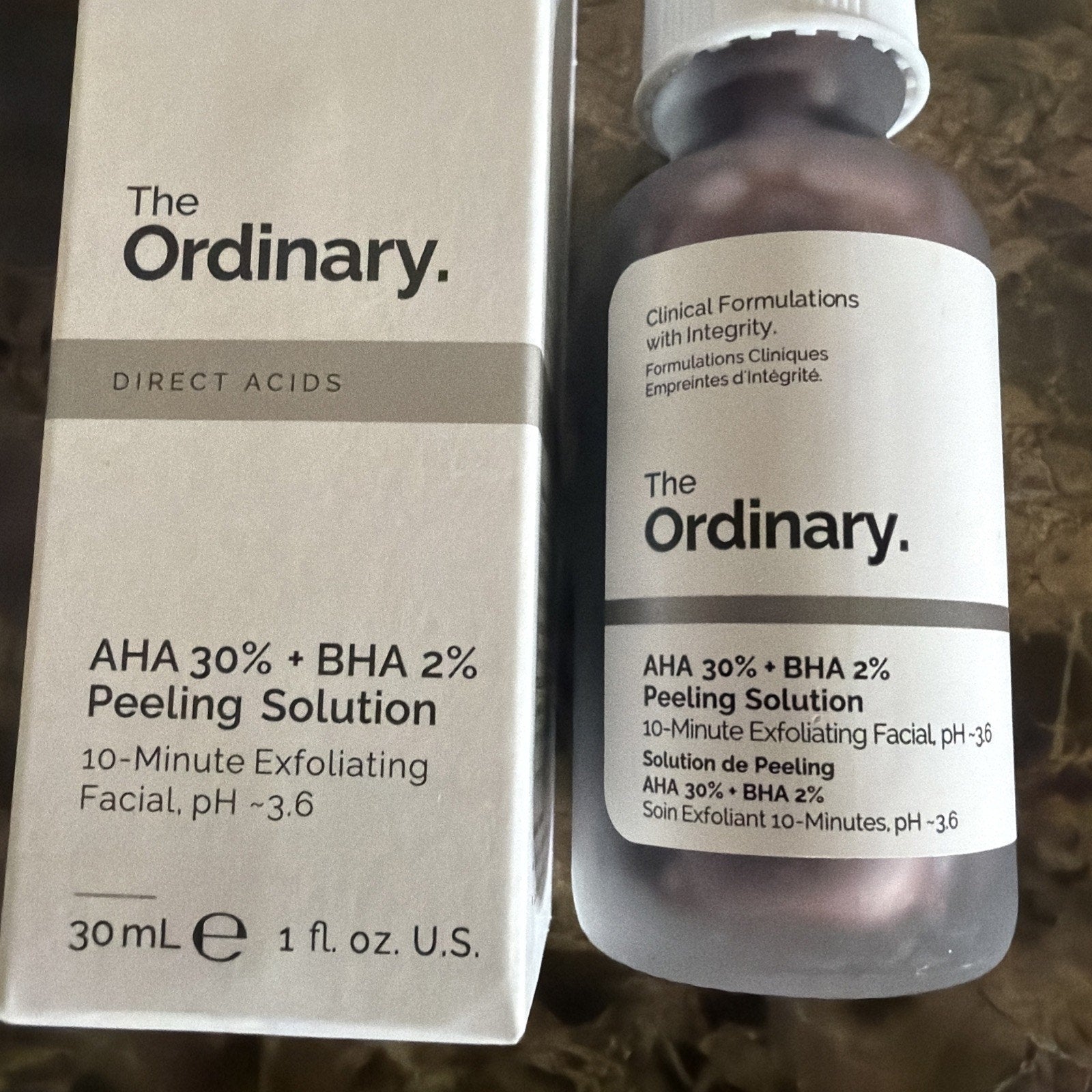 Box and frosted glass bottle of The Ordinary AHA 30% + BHA 2% Peeling Solution with minimalist black text on a white background, placed on a textured surface.