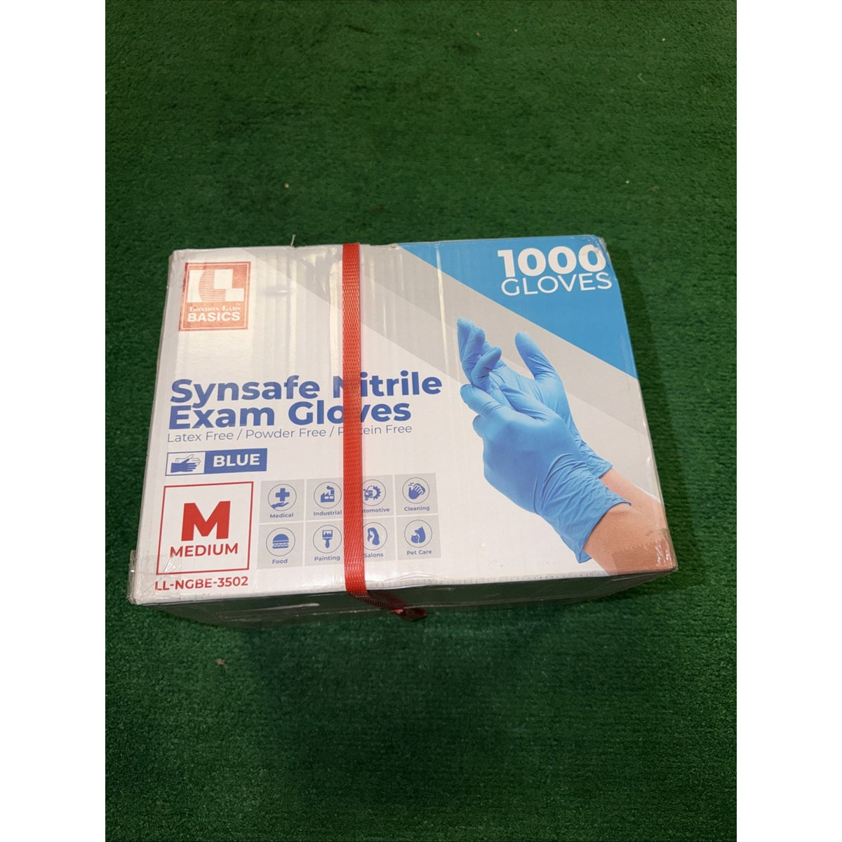 Box containing 1000 medium-sized blue nitrile exam gloves labeled as latex free, powder free, and sterile, with product icons and a red strap securing the box on a green surface.