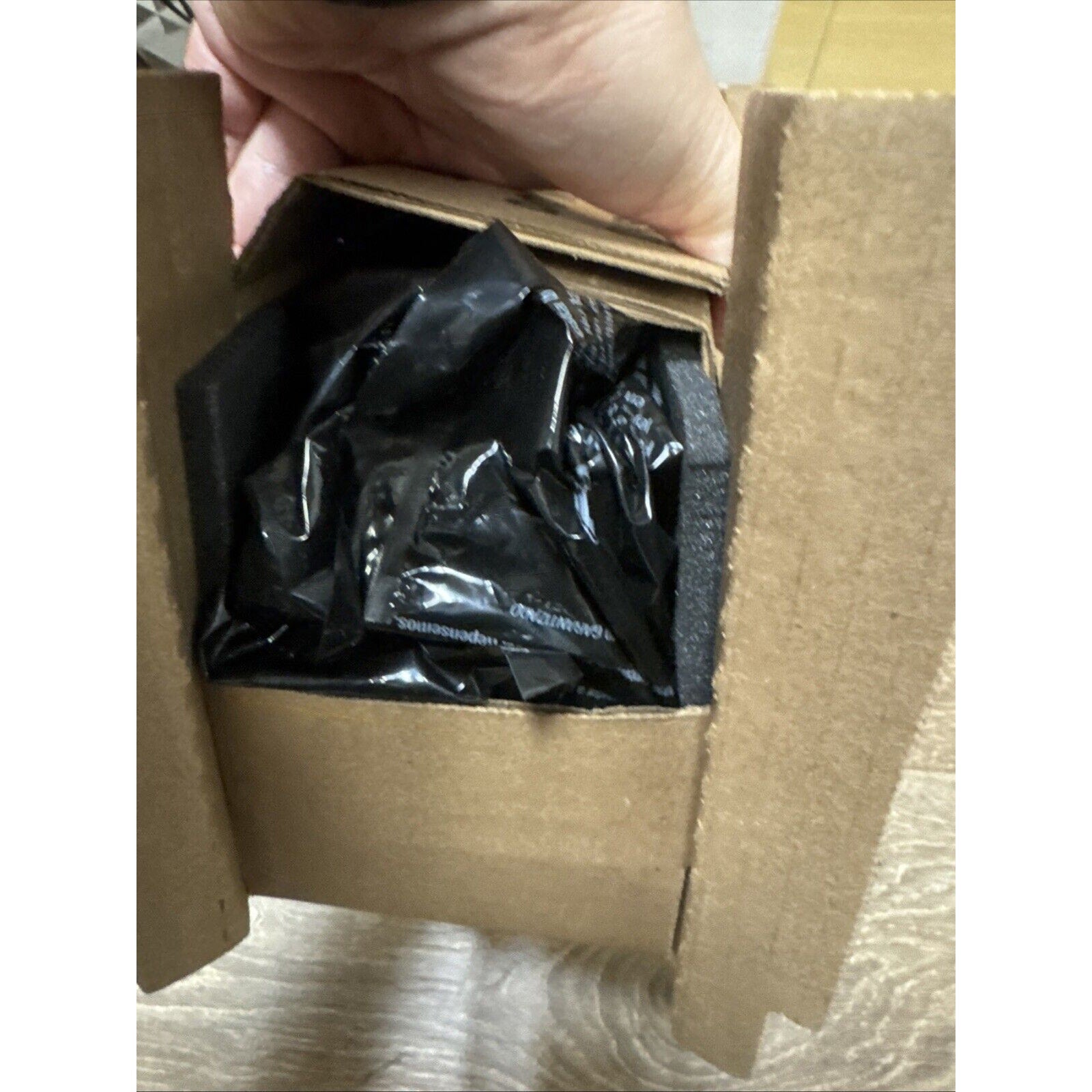 Hand holding open a brown cardboard box revealing a tightly packed black plastic-wrapped item secured by black foam inserts inside the box.