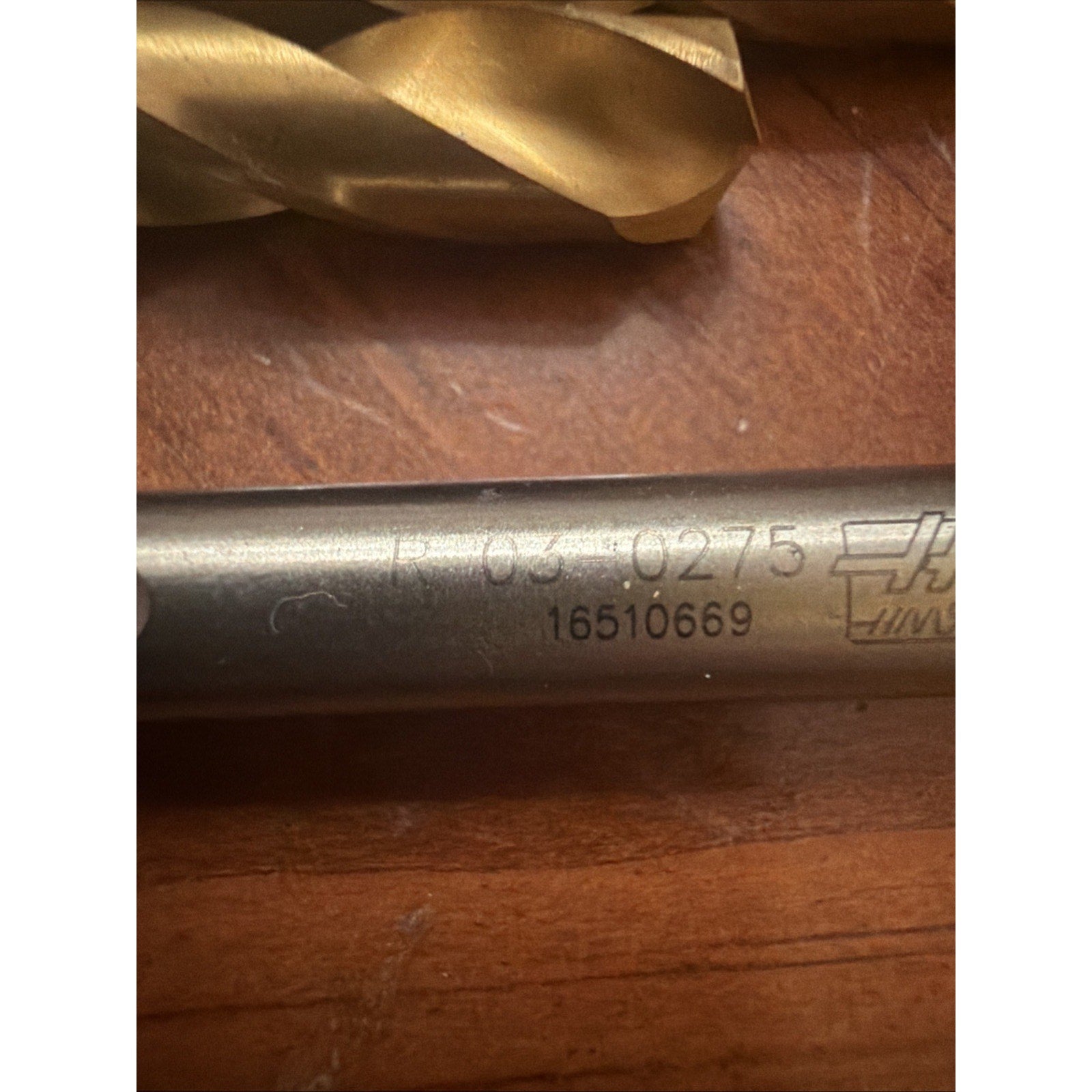 Close-up of a metal tool shaft with engraved identification numbers and part of a gold-colored drill bit visible on a wooden surface.
