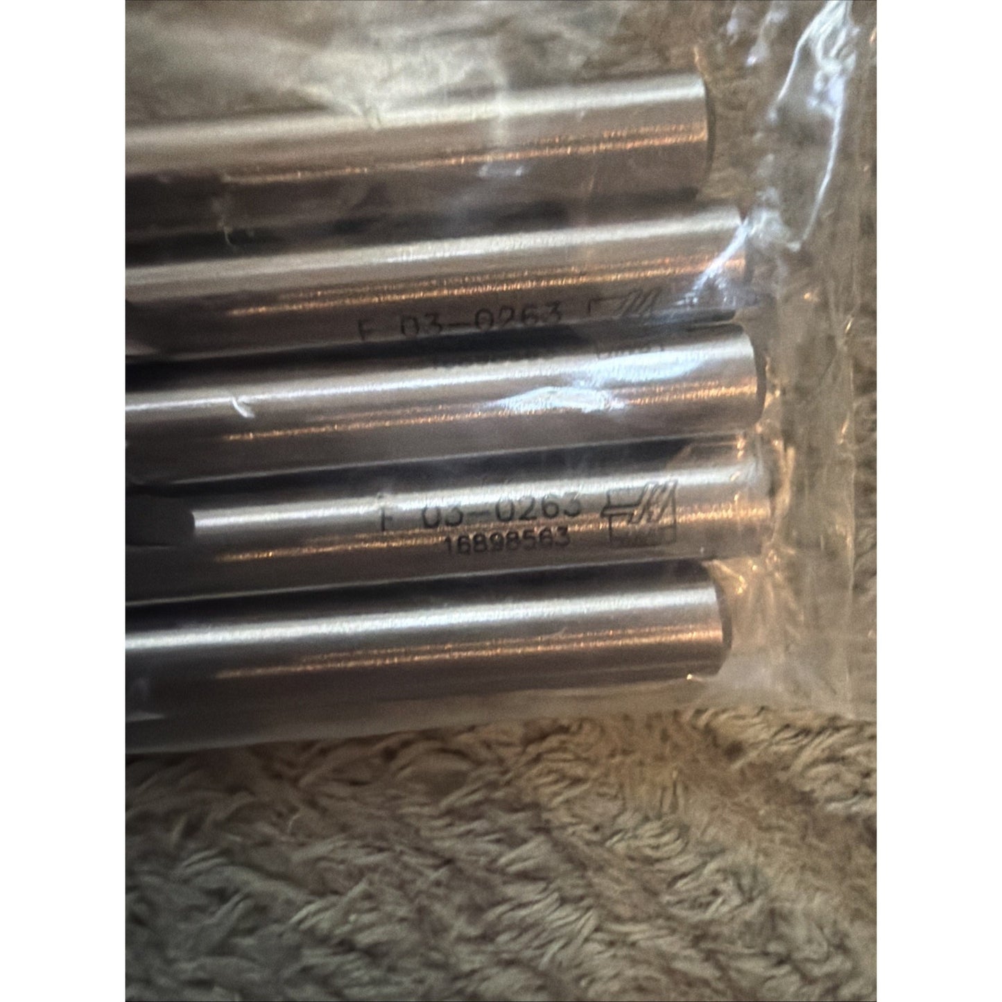 Close-up view of several metallic cylindrical rods with engraved alphanumeric markings, partially enclosed in clear plastic on a textured beige fabric surface.