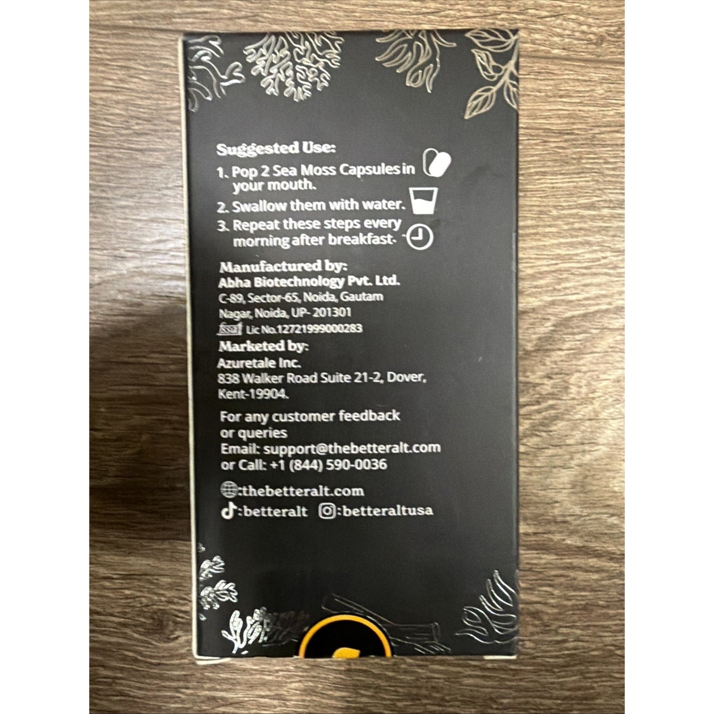 Back of a black dietary supplement box featuring illustrated seaweed designs, usage instructions for sea moss capsules, manufacturer and marketer contact information, and social media handles.