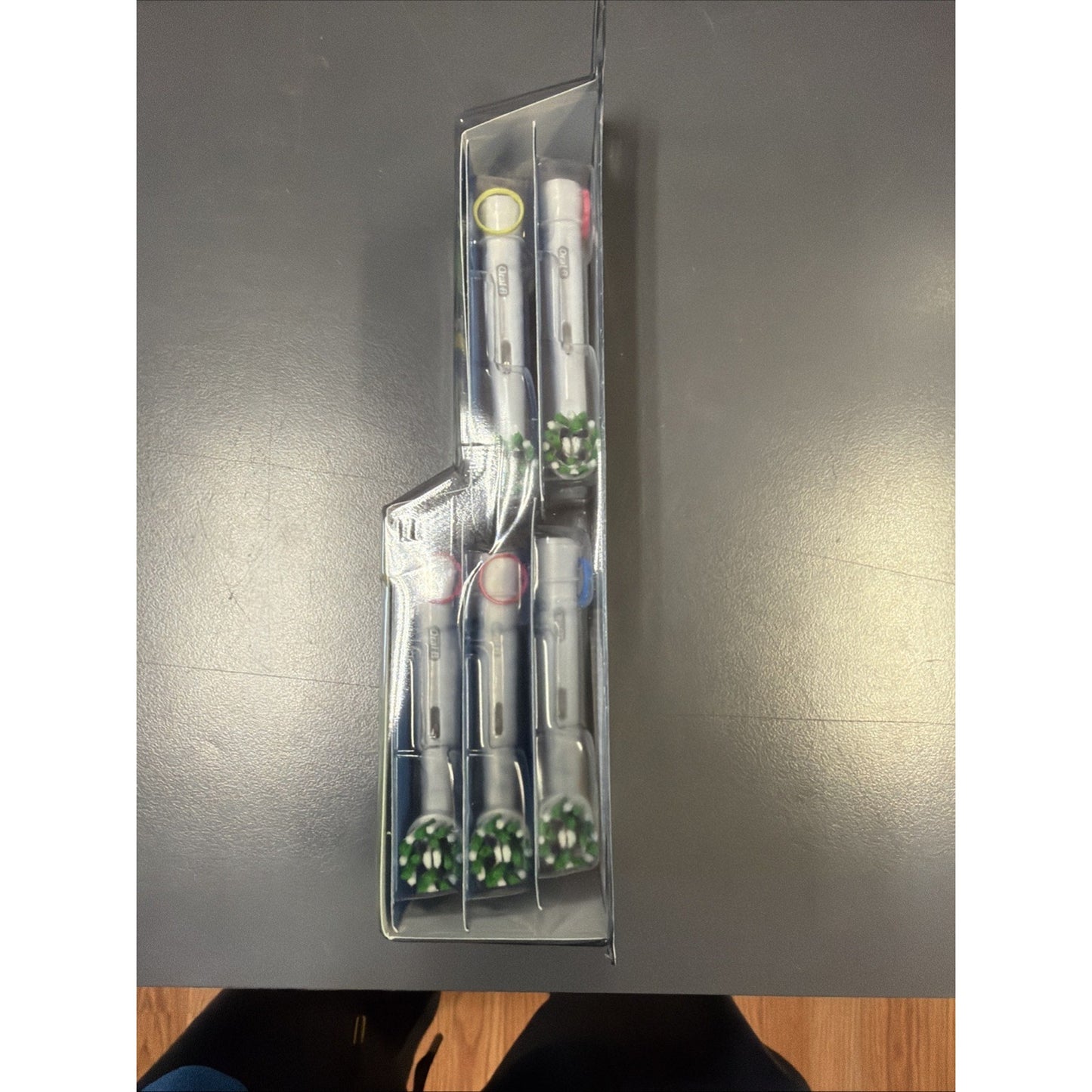 Side view of a clear plastic package containing five electric toothbrush heads with green bristles and colored ring indicators, placed on a dark gray surface.