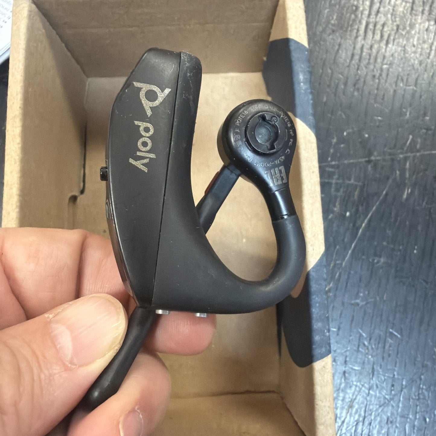 Close-up of a hand holding a black Poly Bluetooth earpiece with an ear hook, inside an open cardboard box on a textured dark surface.