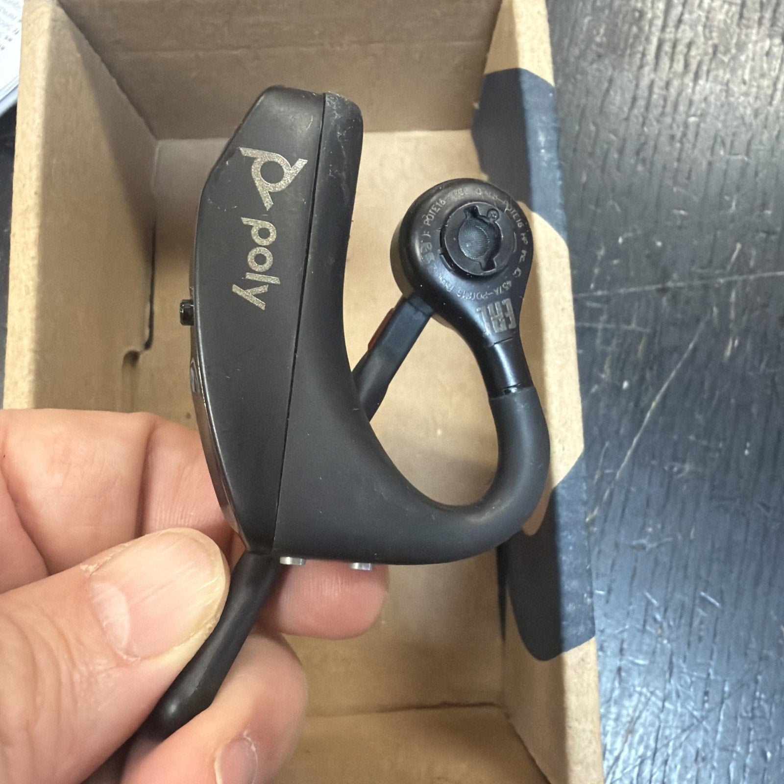 Close-up of a hand holding a black Poly Bluetooth earpiece with an ear hook, inside an open cardboard box on a textured dark surface.
