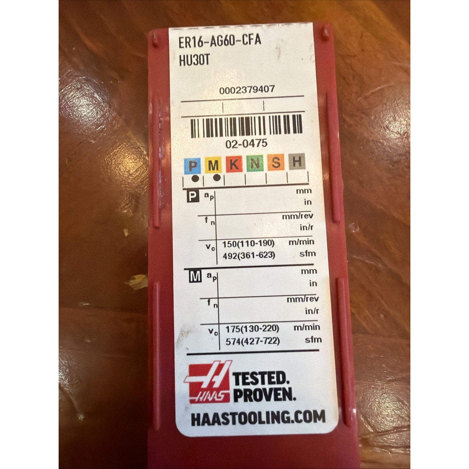 Narrow red plastic case with a white label showing colored metal type codes, detailed machining parameters, a barcode, and the Haas logo with the text "Tested. Proven." on a wooden surface.