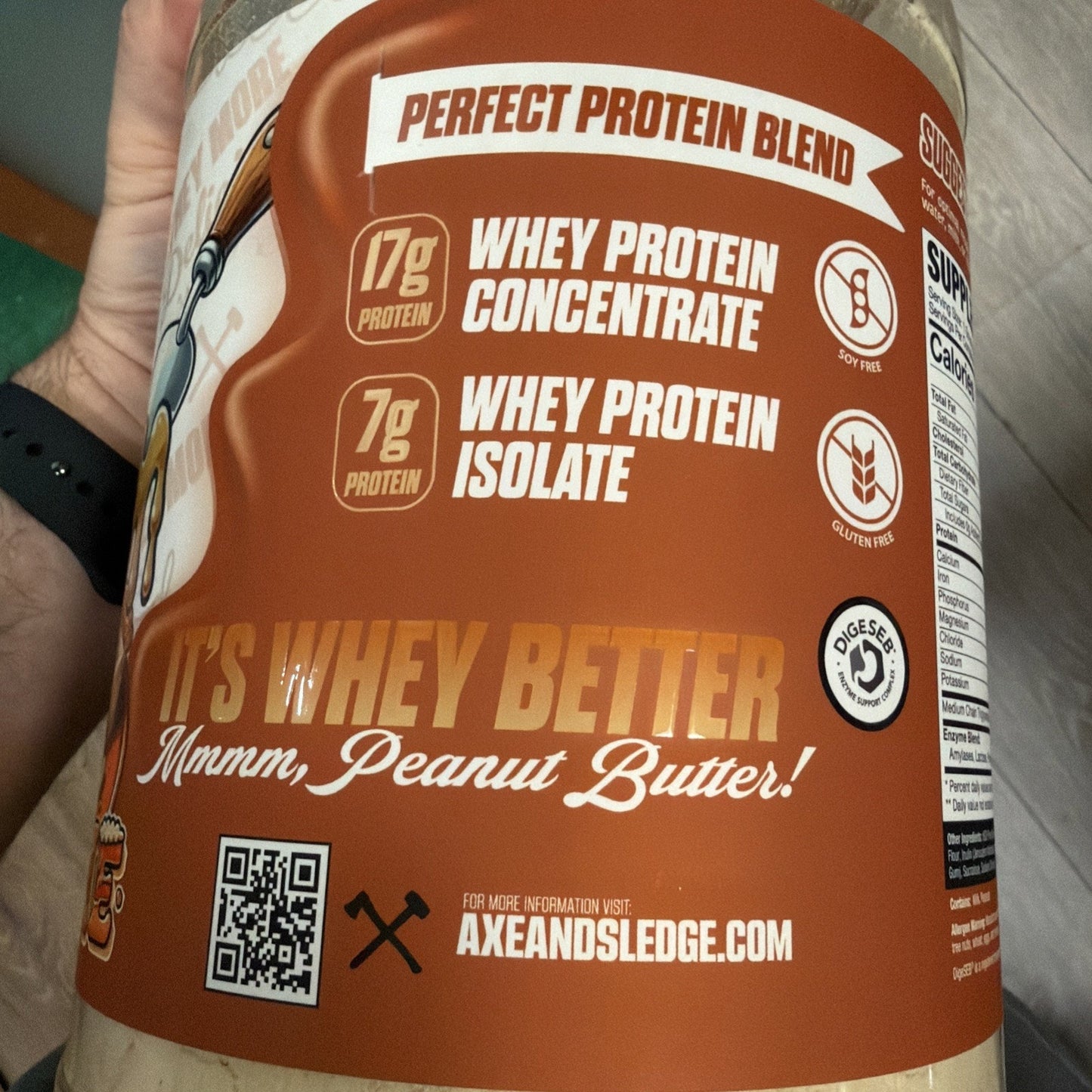 Close-up of a peanut butter-flavored protein powder container label highlighting 17g whey protein concentrate, 7g whey protein isolate, soy-free, gluten-free, and a QR code for more information.