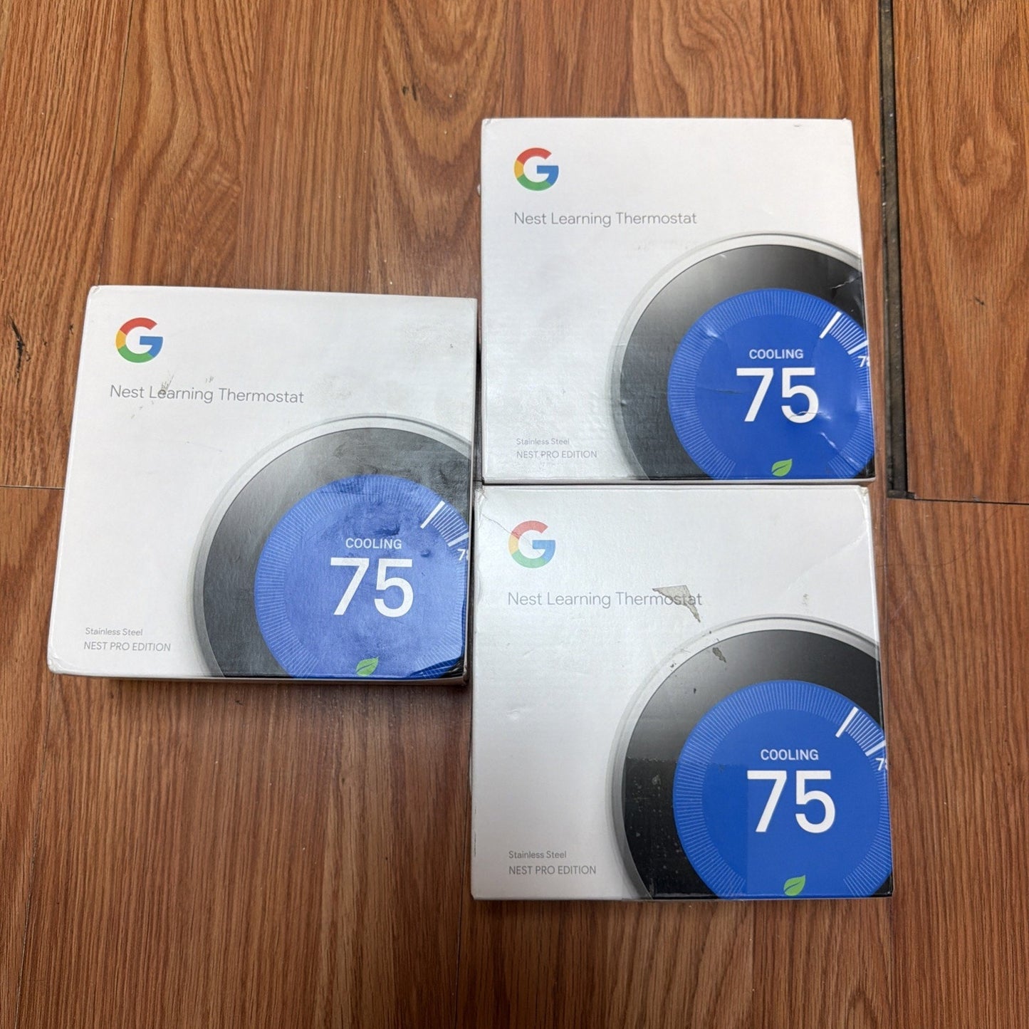 Three boxed Google Nest Learning Thermostats displayed on a wooden floor, each showing a close-up image of the thermostat dial set to cooling mode at 75 degrees.