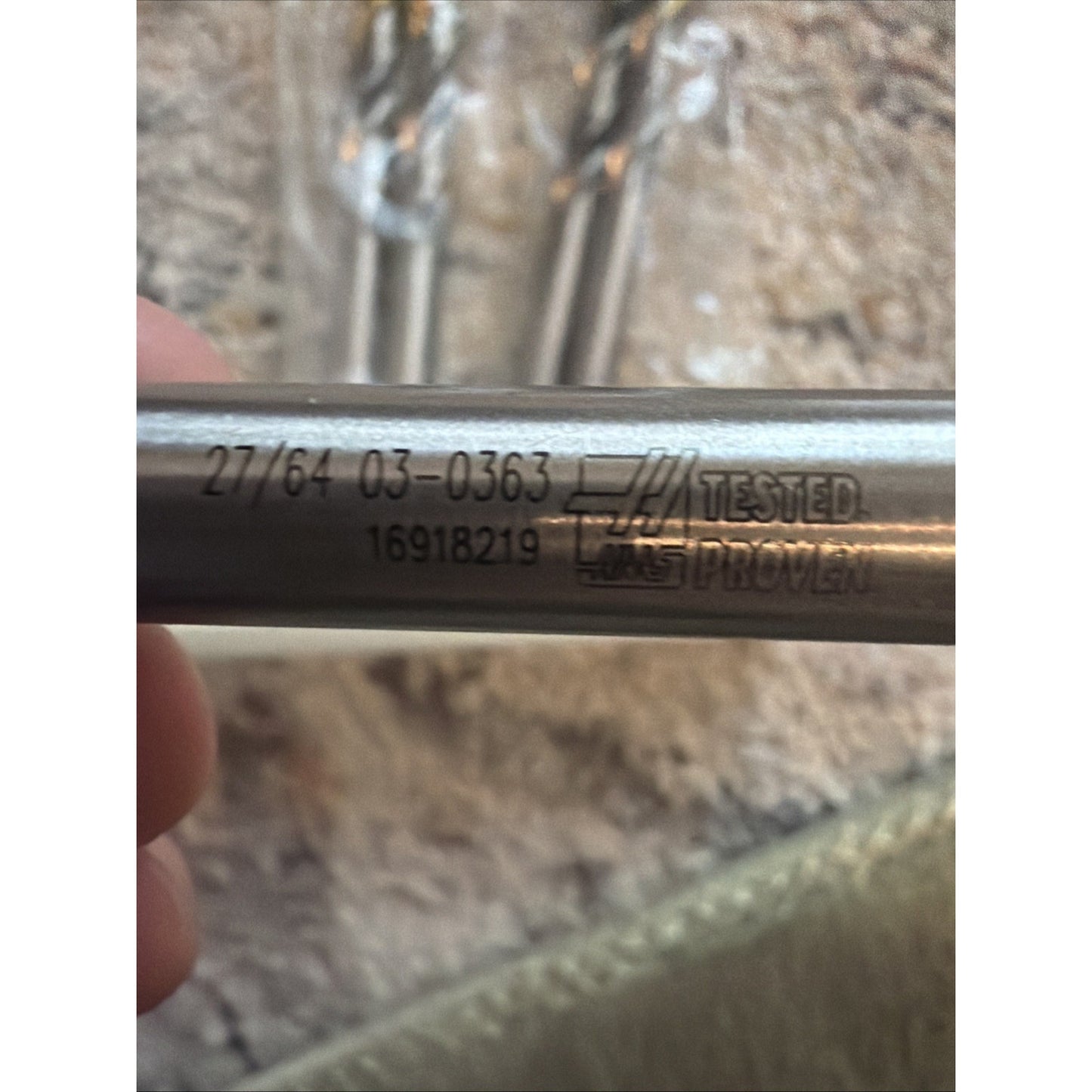 Close-up of a silver cylindrical metal rod with engraved numbers and a "Tested Proven" logo, held by a hand with blurred similar rods and a textured light surface in the background.