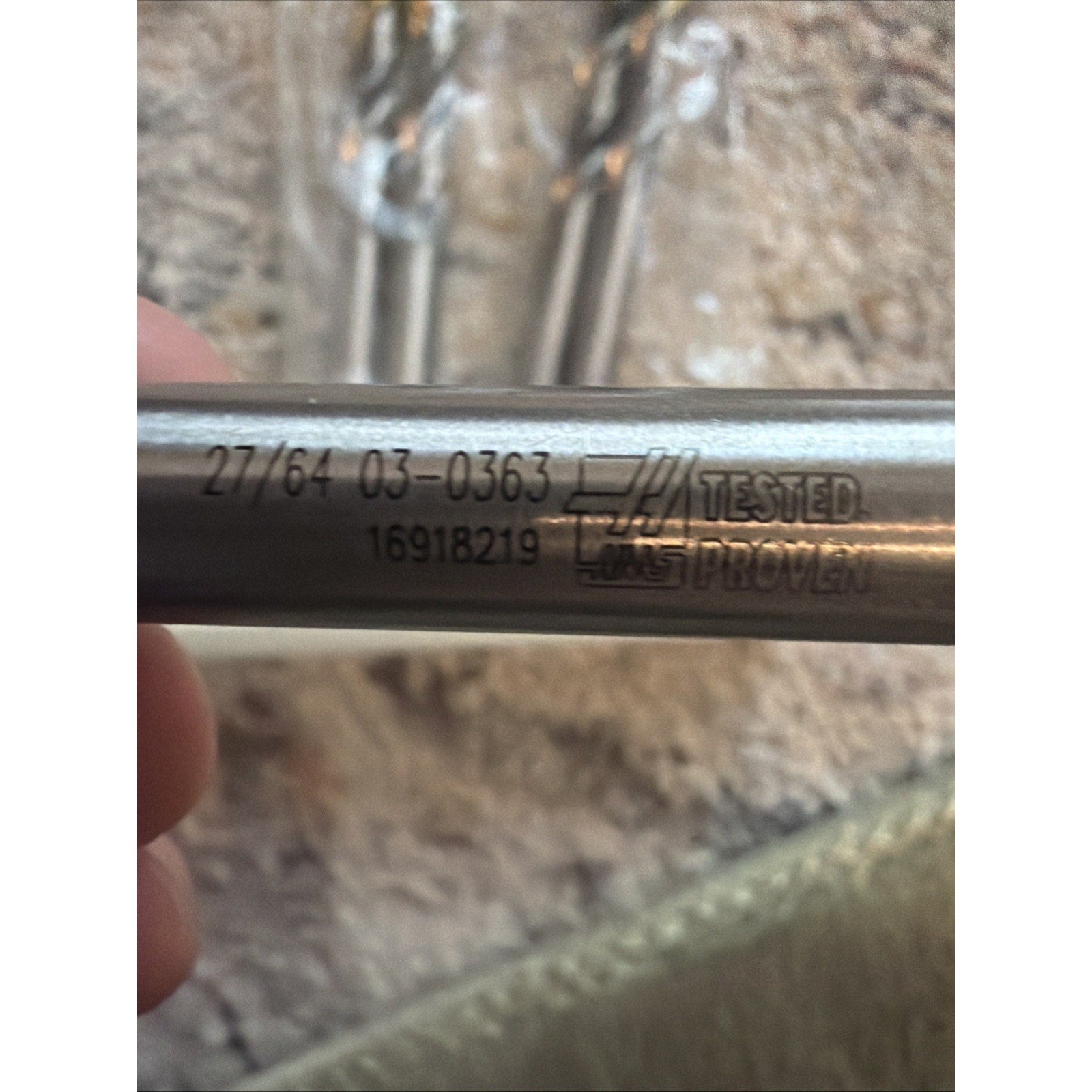 Close-up of a silver cylindrical metal rod with engraved numbers and a "Tested Proven" logo, held by a hand with blurred similar rods and a textured light surface in the background.