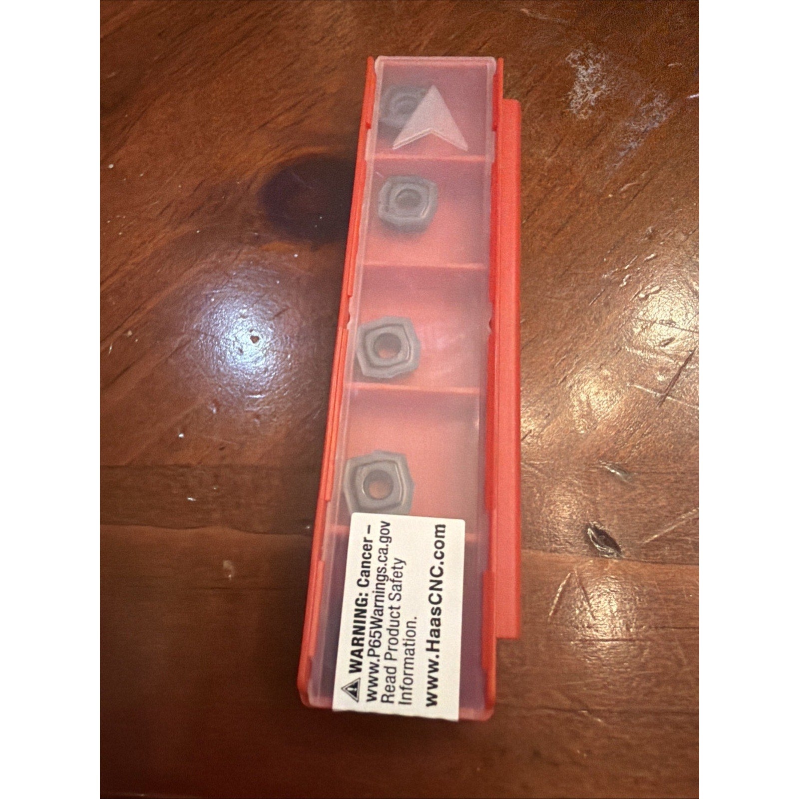 Red plastic case with a translucent lid containing four metal cutting inserts in separate compartments, placed on a wooden surface with a visible warning label on the case.