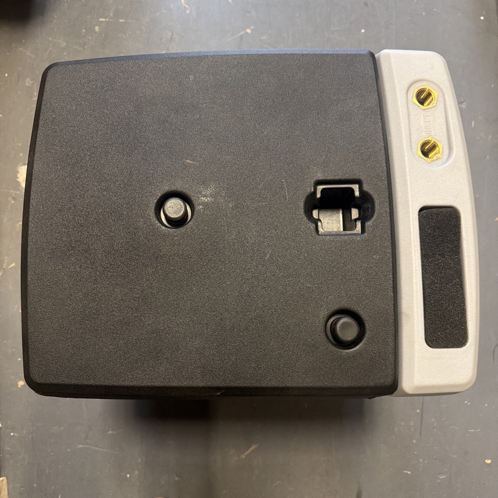 Top-down view of a black rectangular device with a gray handle and two brass connectors on the right side, featuring two round buttons and a latch on the black surface.
