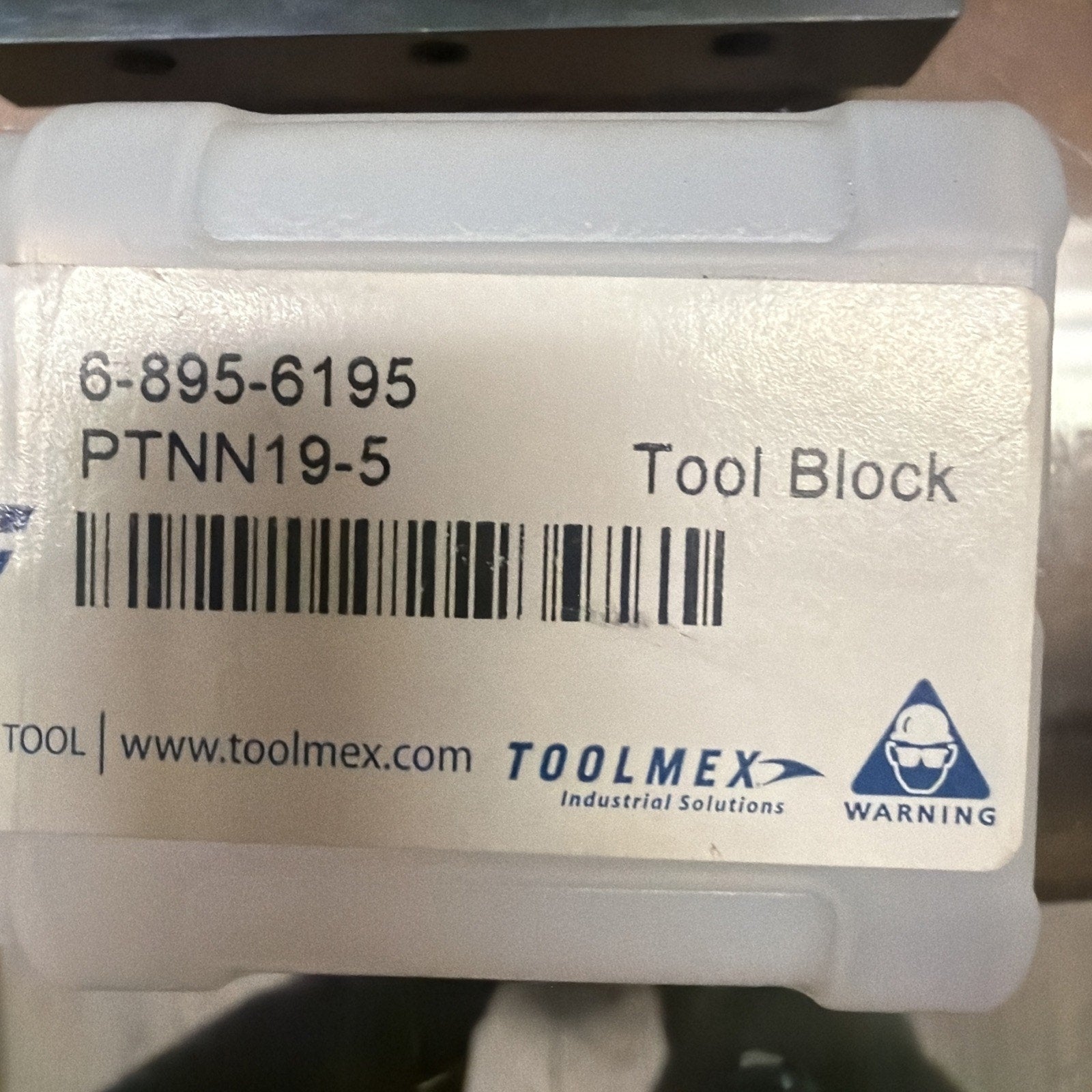 White label on a translucent plastic container displaying product code "6-895-6195," model "PTNN19-5," the words "Tool Block," a barcode, the Toolmex logo with the text "Industrial Solutions," a website URL, and a blue warning icon.