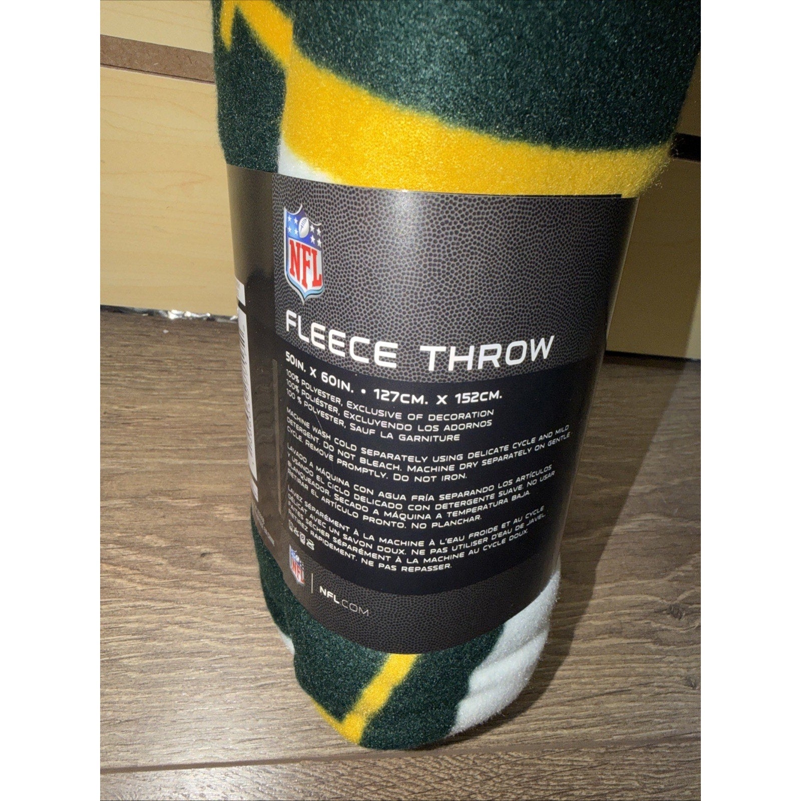 Rolled fleece throw blanket wrapped with a black label featuring the NFL logo, product dimensions, and care instructions in English and Spanish, resting on a wooden floor.