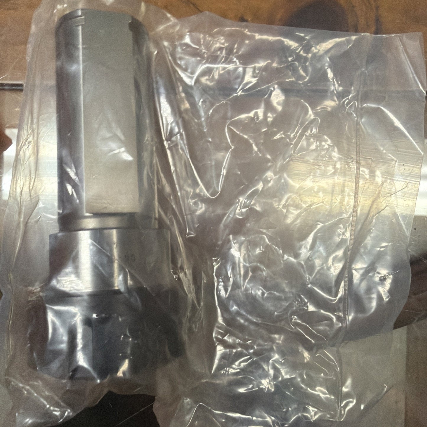 Metallic mechanical component with a cylindrical body and hexagonal section, partially wrapped in a transparent plastic bag, resting on a wooden surface.