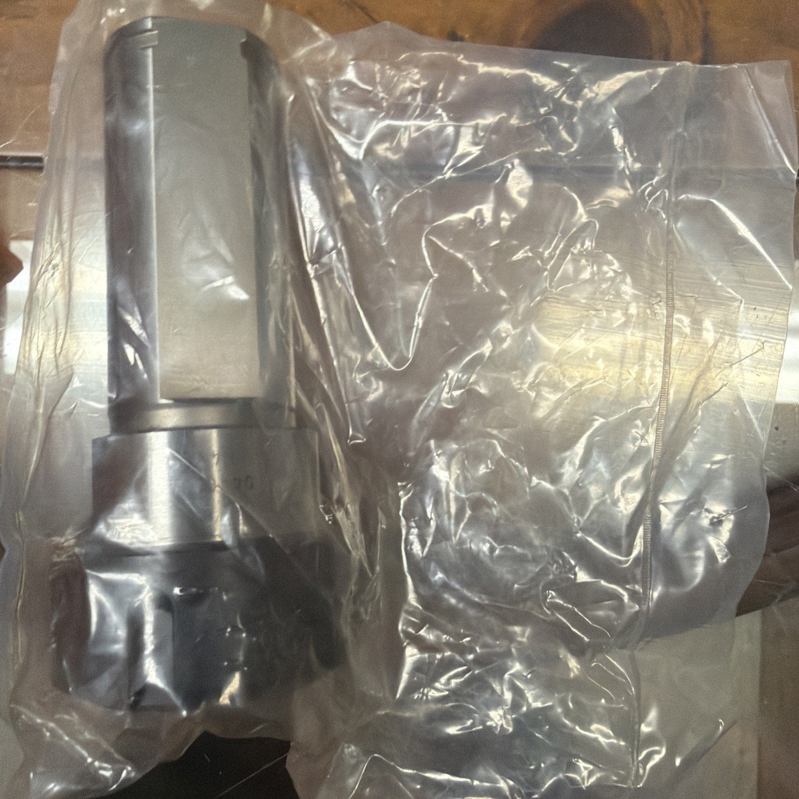 Metallic mechanical component with a cylindrical body and hexagonal section, partially wrapped in a transparent plastic bag, resting on a wooden surface.