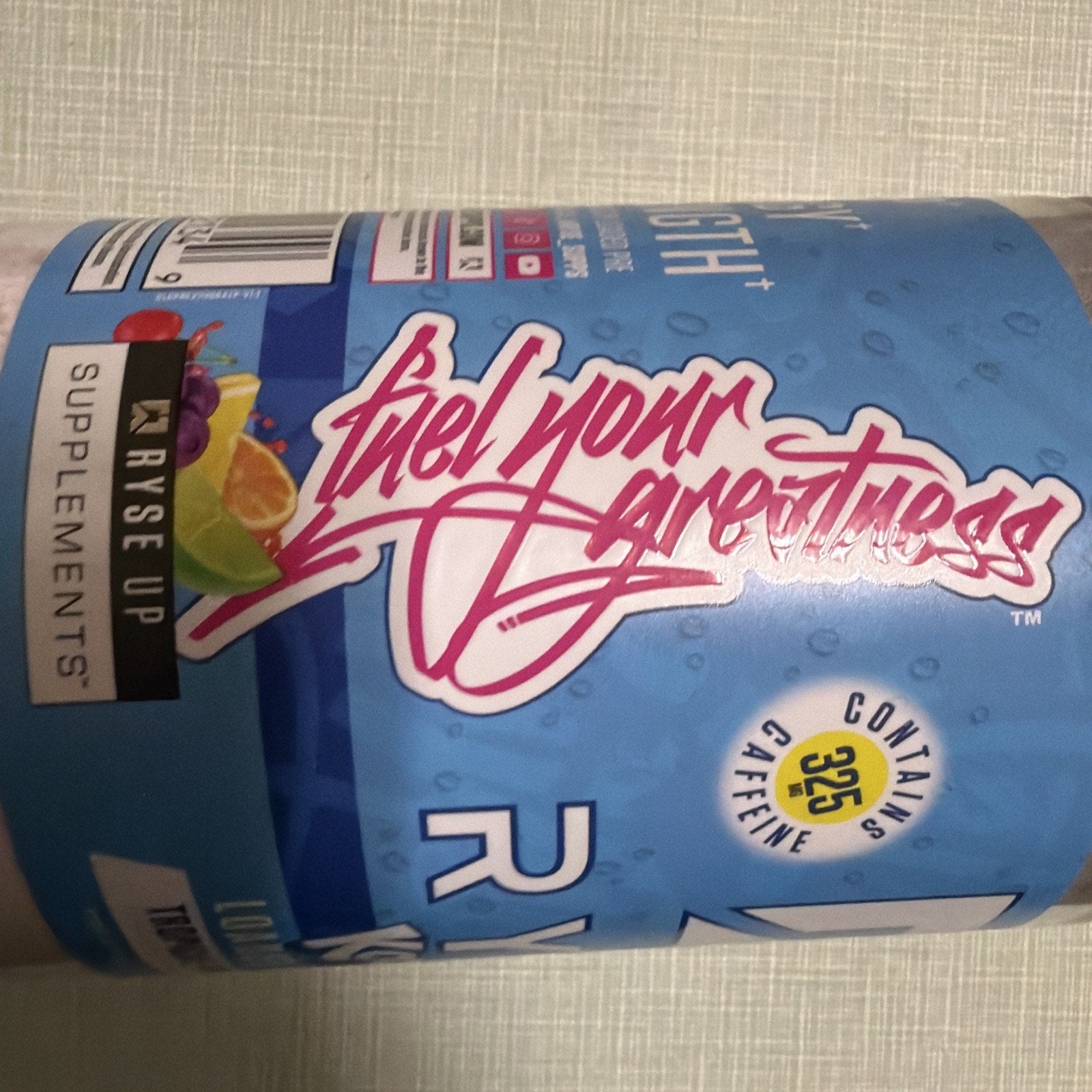 Close-up of a blue supplement container label featuring the slogan "Fuel your greatness" in stylized pink script, with icons indicating 325 mg caffeine content and branding for a supplement company.