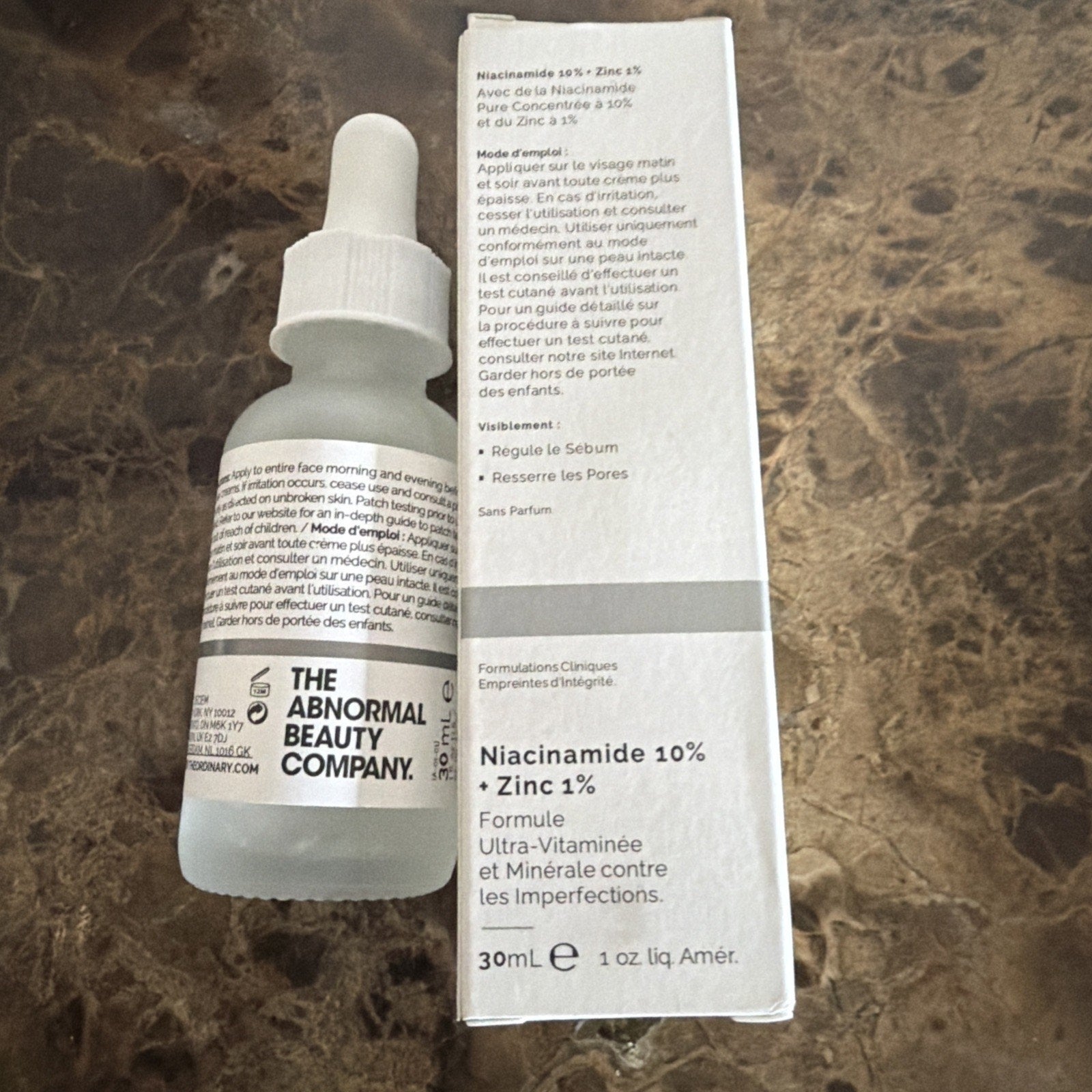 Clear frosted glass dropper bottle labeled The Abnormal Beauty Company with adjacent white box detailing a Niacinamide 10% and Zinc 1% formula for skin with usage instructions in French on a dark marble surface.