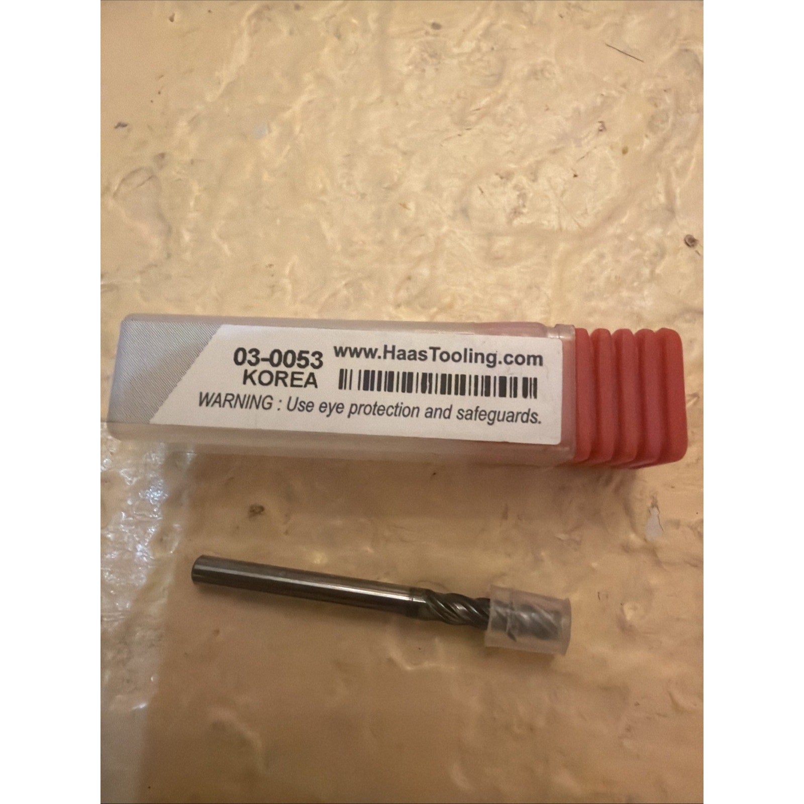 Metal cutting tool bit with a short spiral flute and protective clear plastic cap, placed beside a clear plastic case with a white label and red end cap on a textured beige surface.