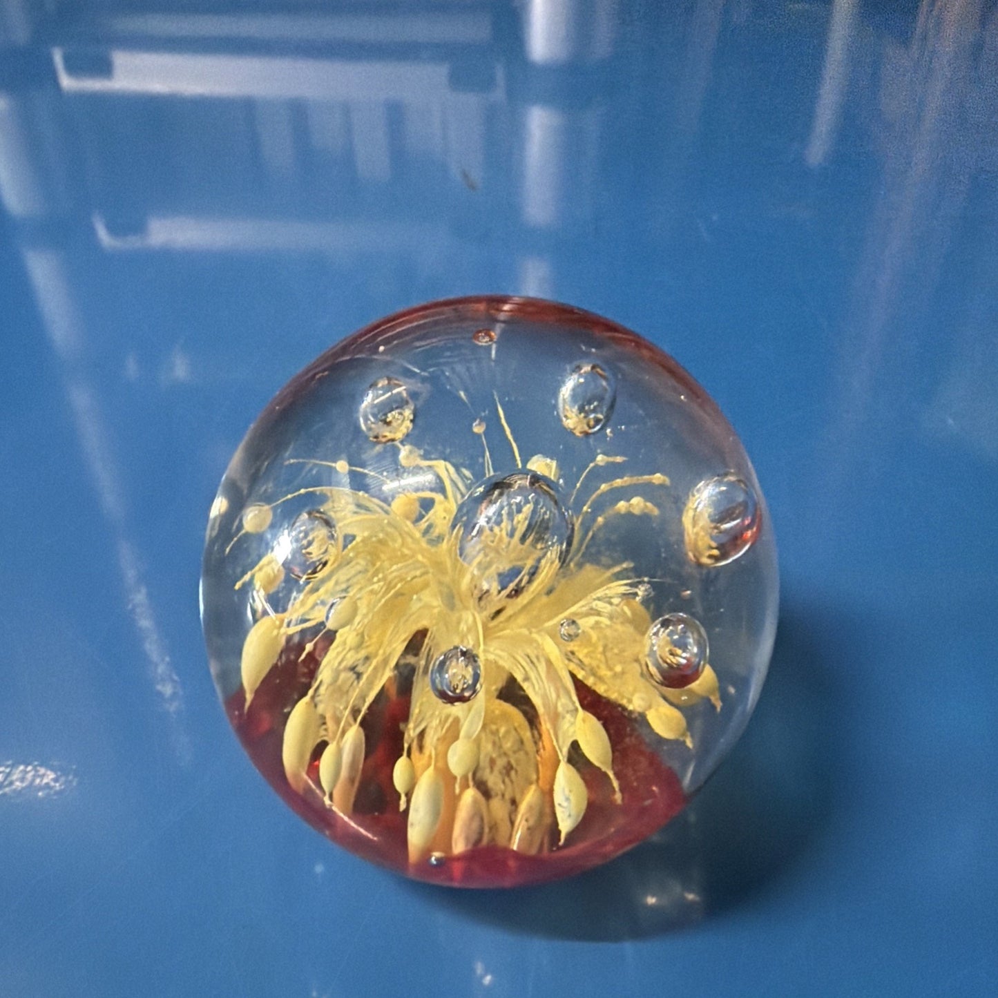 Clear glass paperweight with a yellow floral design and embedded clear bubbles, placed on a glossy blue surface with reflections visible.