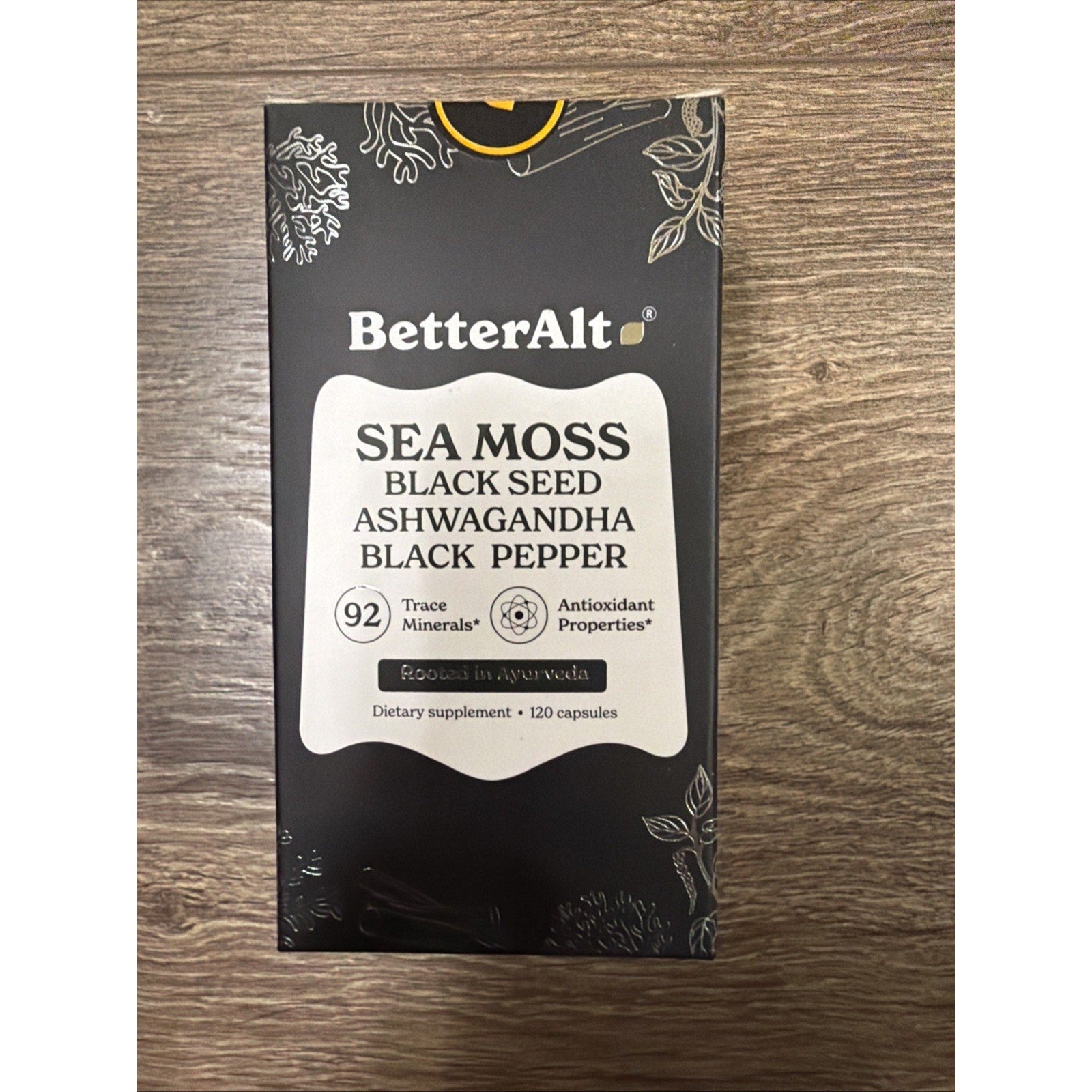 Rectangular black box packaging for BetterAlt dietary supplement featuring sea moss, black seed, ashwagandha, and black pepper, listing 92 trace minerals and antioxidant properties on a wooden surface.