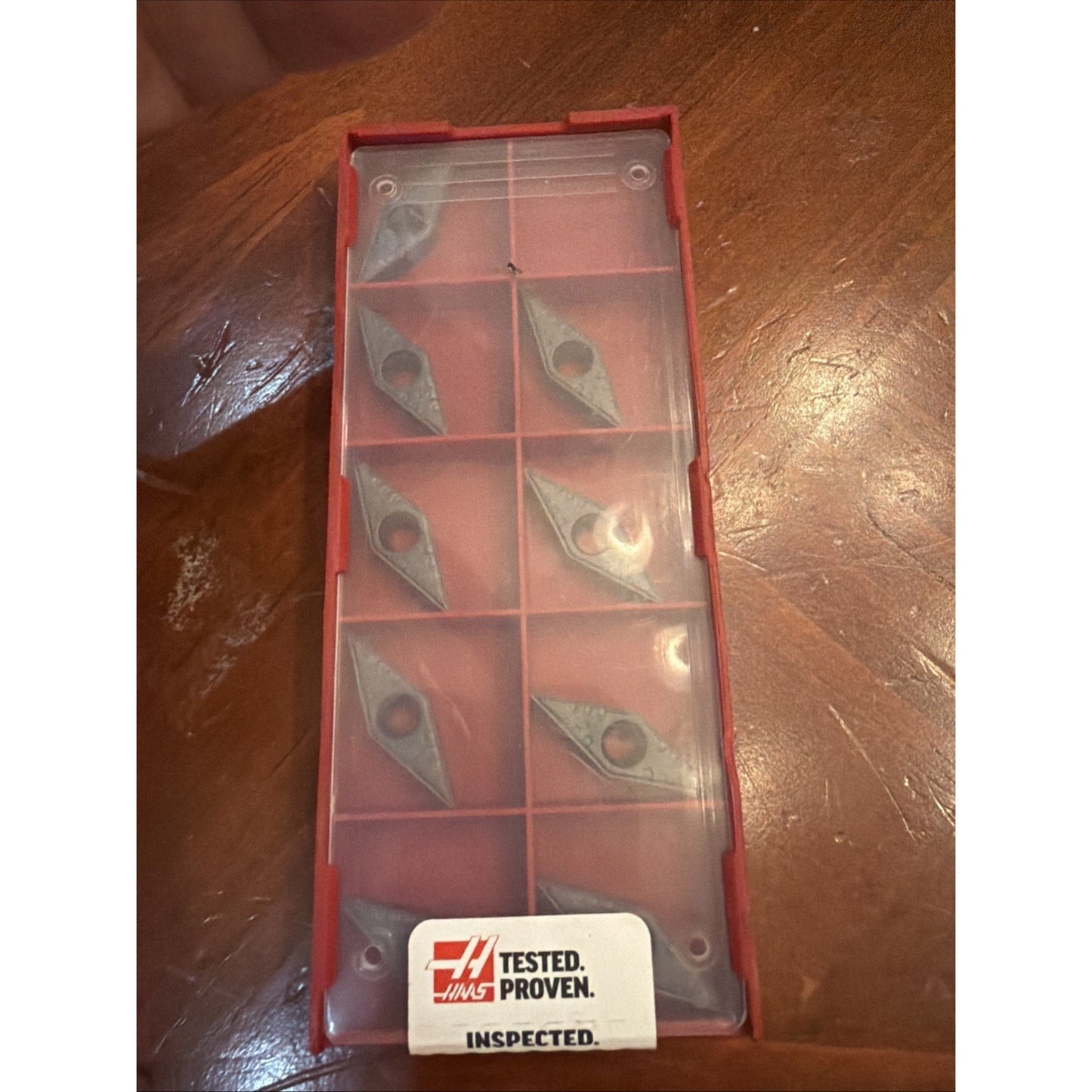 Red rectangular plastic case with a translucent lid containing eight gray diamond-shaped metal inserts arranged in individual compartments, placed on a wooden surface with a Haas Tested Proven Inspected sticker.