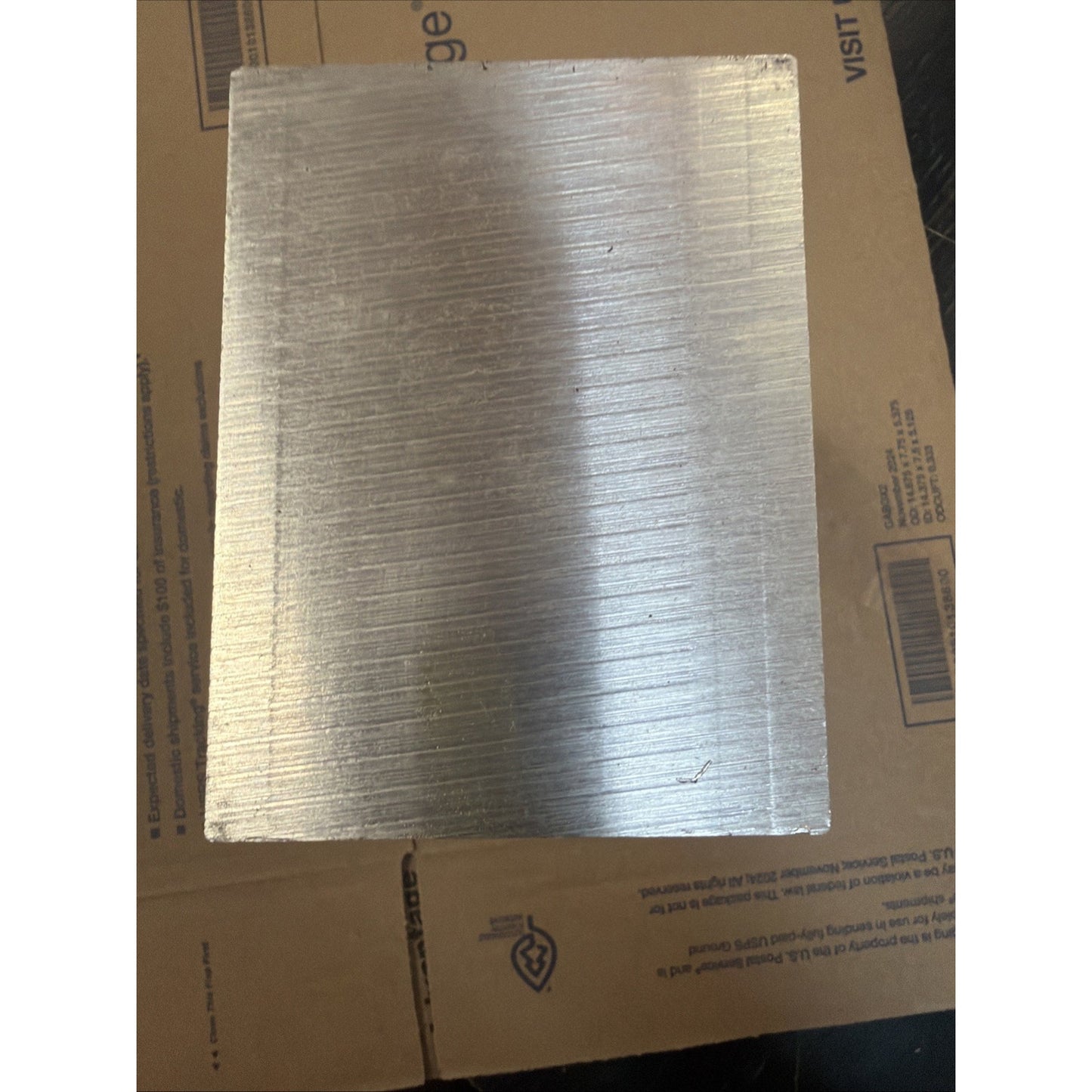 Square metal plate with a brushed texture showing fine horizontal lines, resting on a cardboard surface with printed text and symbols partially visible.