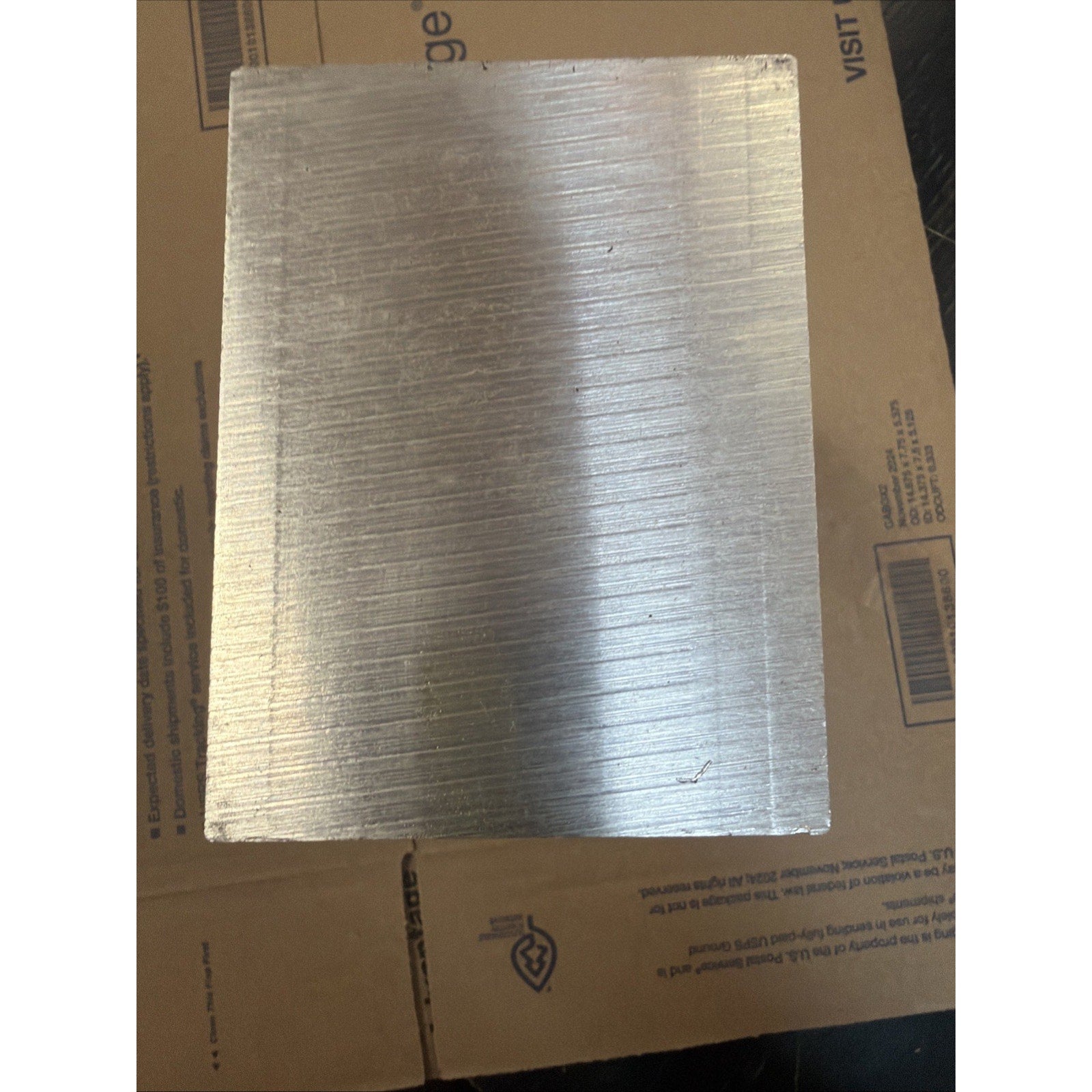 Square metal plate with a brushed texture showing fine horizontal lines, resting on a cardboard surface with printed text and symbols partially visible.