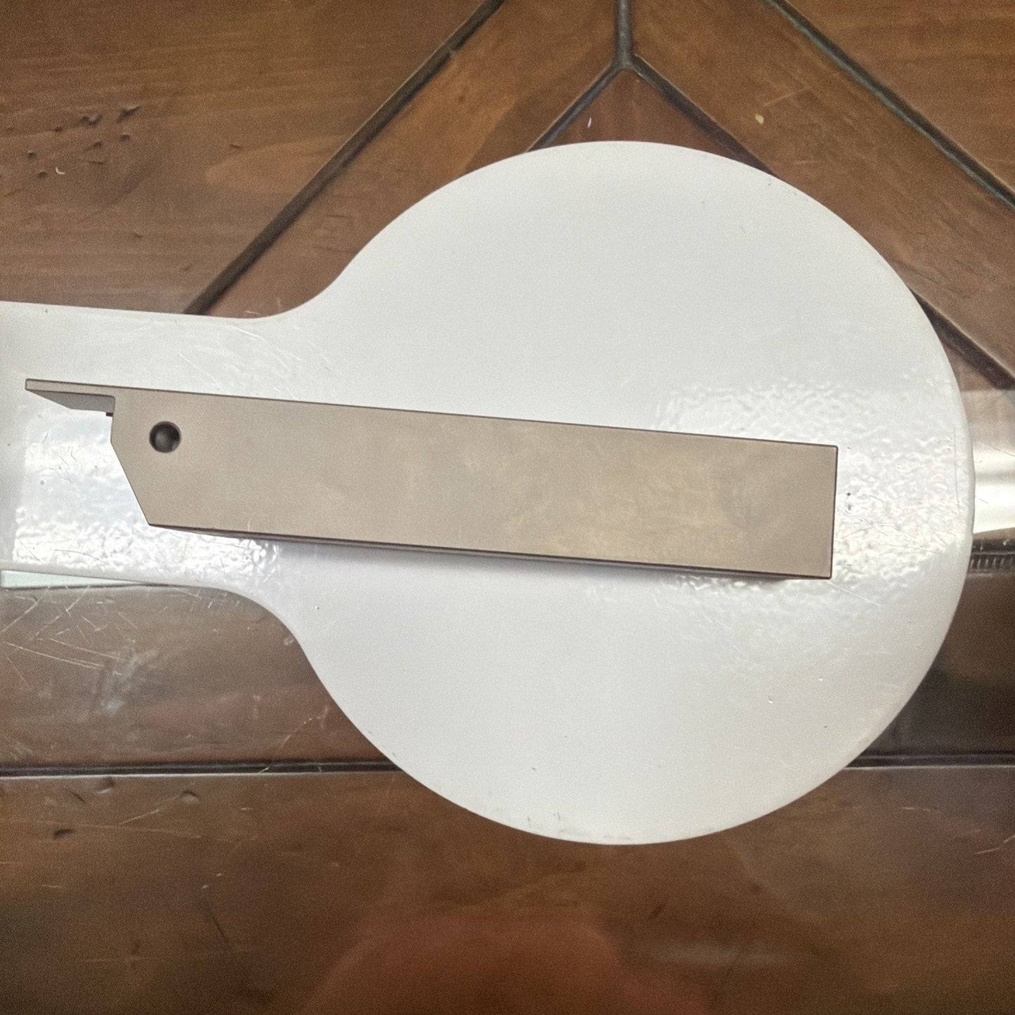 Rectangular metal lathe cutting tool with a small hole near one end, resting on a round white surface over a wooden table with visible grain and seams.