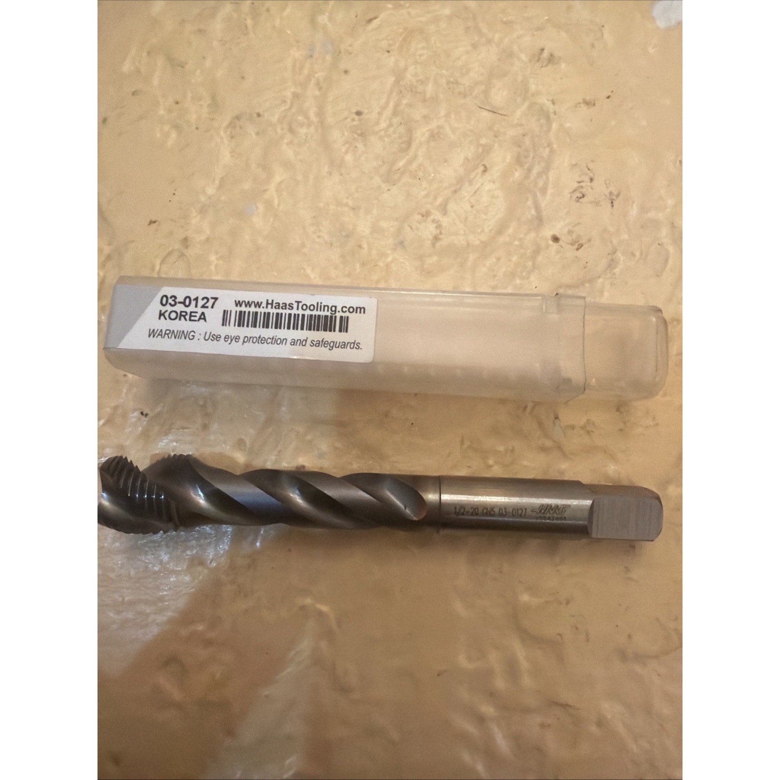 Spiral-fluted metal threading tap with a square drive shank, resting on a textured beige surface next to a clear plastic storage tube labeled with a barcode and safety warning.