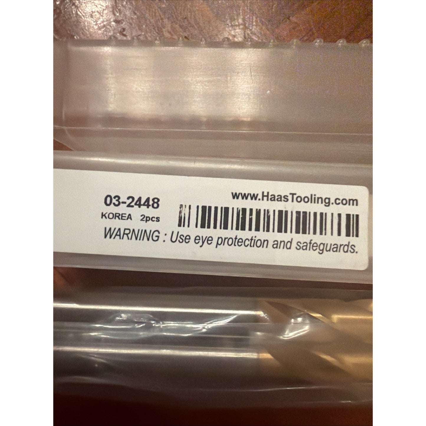 Close-up of a white label on a clear plastic case showing product code 03-2448, origin marked as Korea, a barcode, website URL, and a safety warning to use eye protection and safeguards.