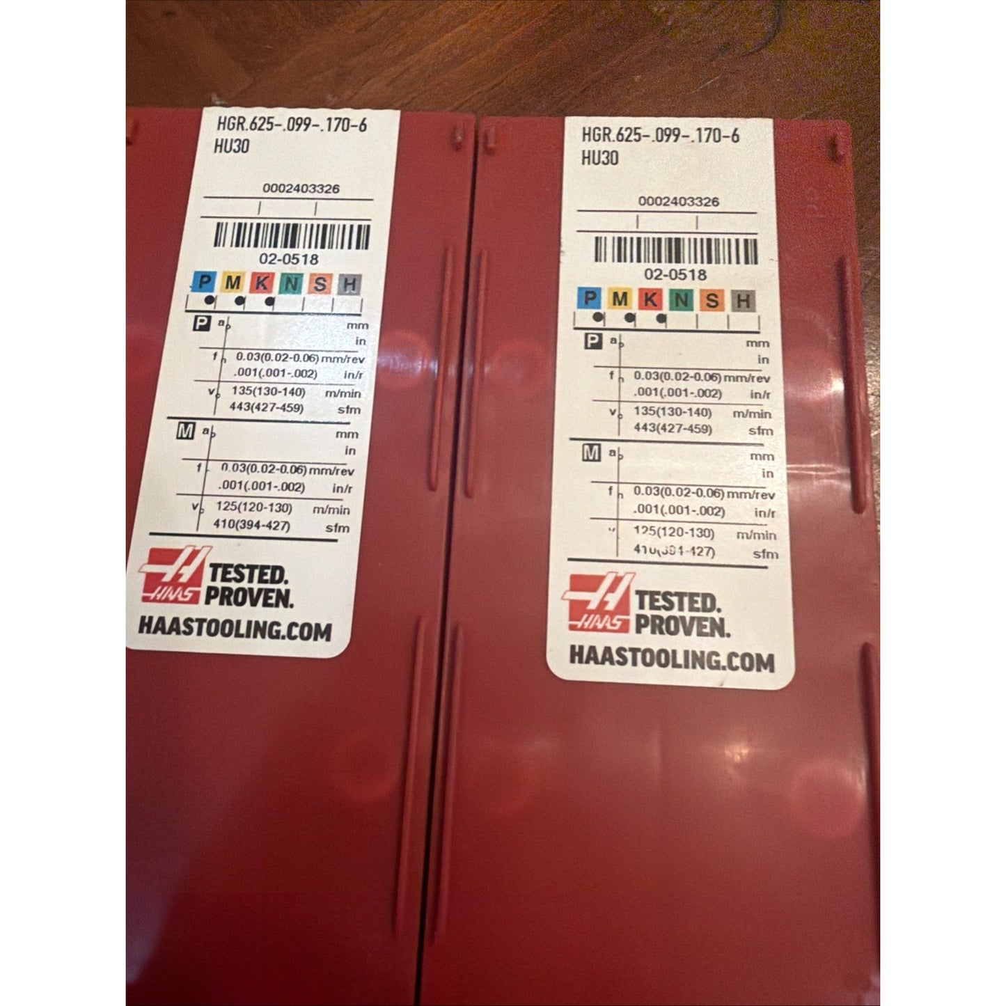 Two red plastic rectangular containers with white labels showing product code "HGR 625-099-170-6 HU30," barcodes, machining parameters, Haas logo, and website, placed on a wooden surface.
